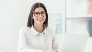 A young woman wearing glasses seated at a computer in a bright office
